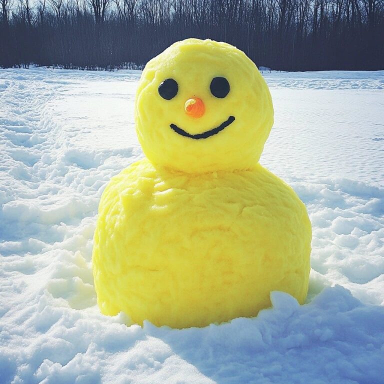A bright yellow two-tiered snow figure with black round eyes, a small orange nose, and a curved black mouth sits on white snow in an open field with bare trees along the distant treeline.